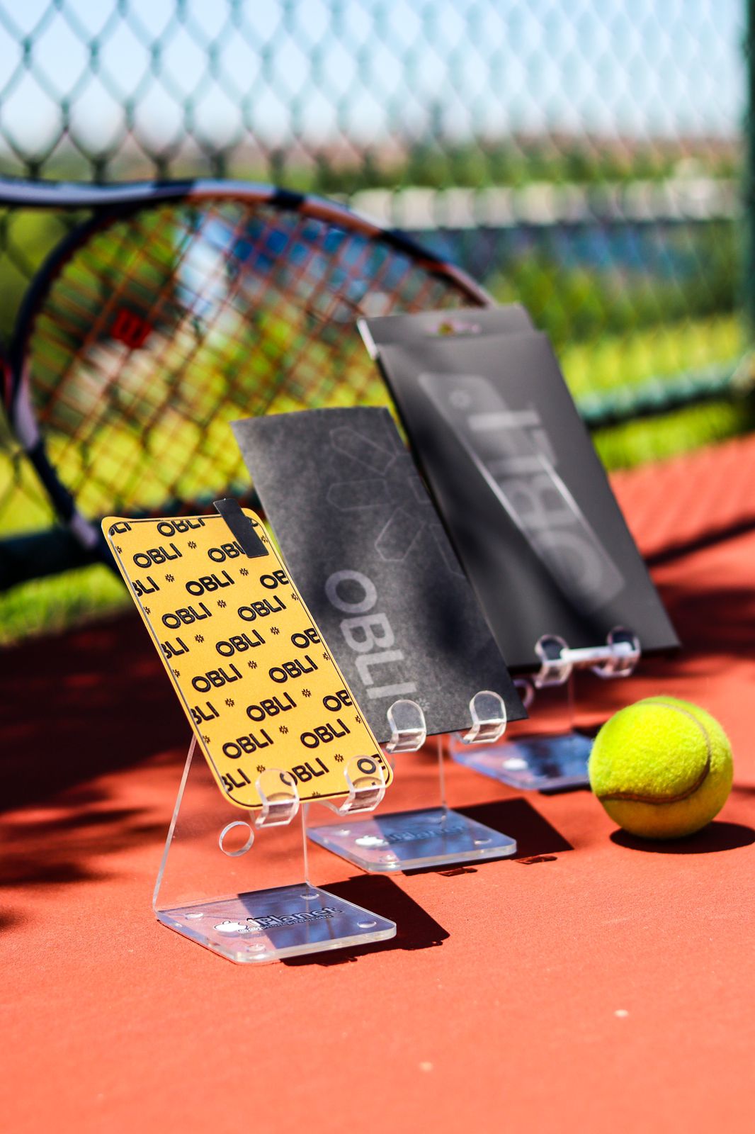 Obli sports accessories and patterned phone case photographed beside a tennis racket and ball on a red court.