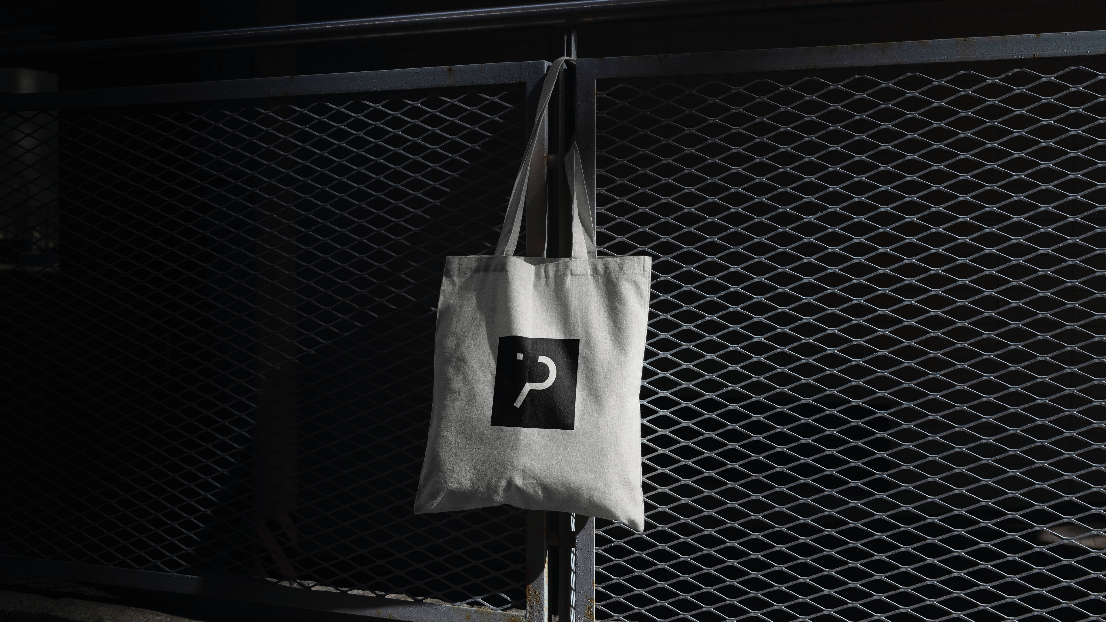 iPlanet fabric tote bag with the black brand symbol hanging on a dark metal fence.
