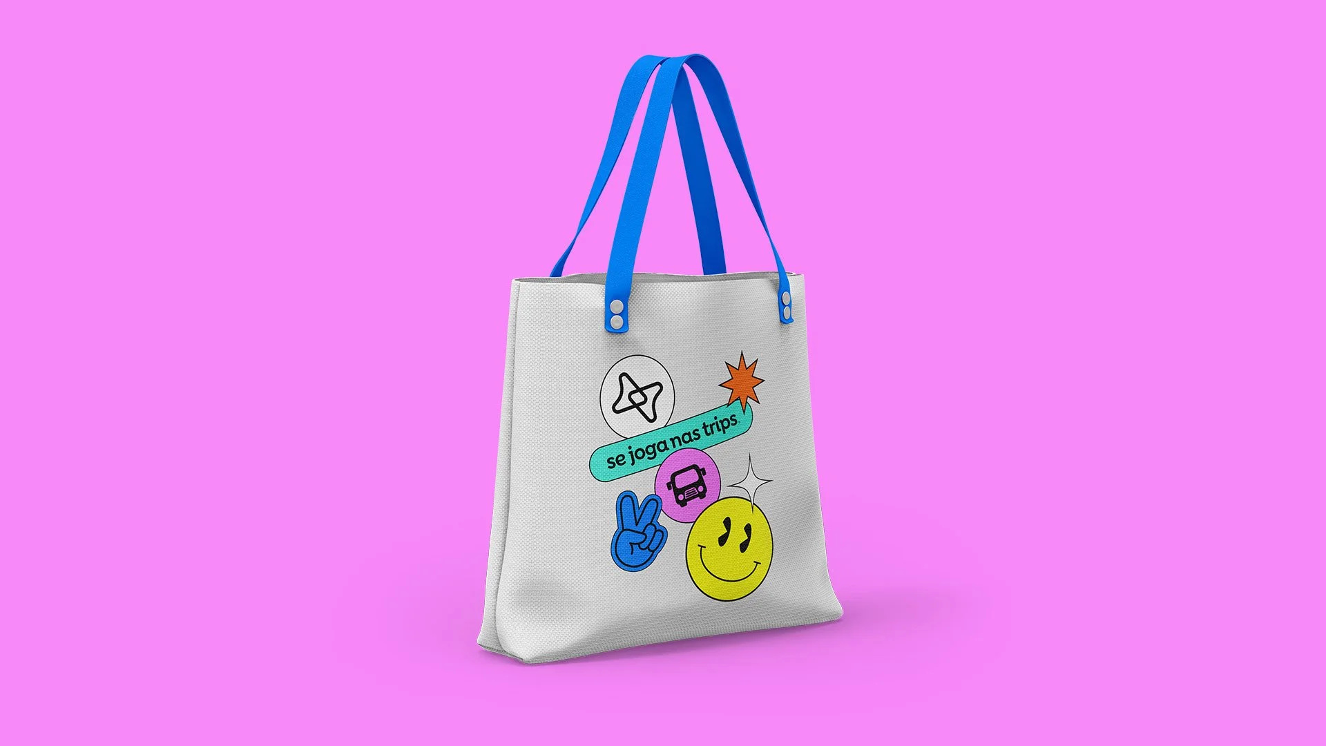 Se Joga Nas Trips white tote bag with blue straps and colorful travel stickers on a pink background.