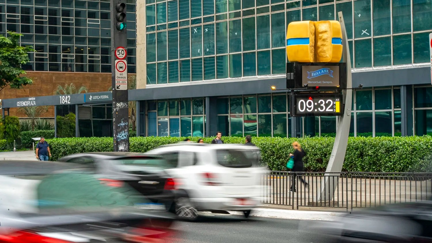 Faixa Azul outdoor campaign with a giant cheese wheel on a street clock, seen behind passing traffic on a São Paulo avenue.